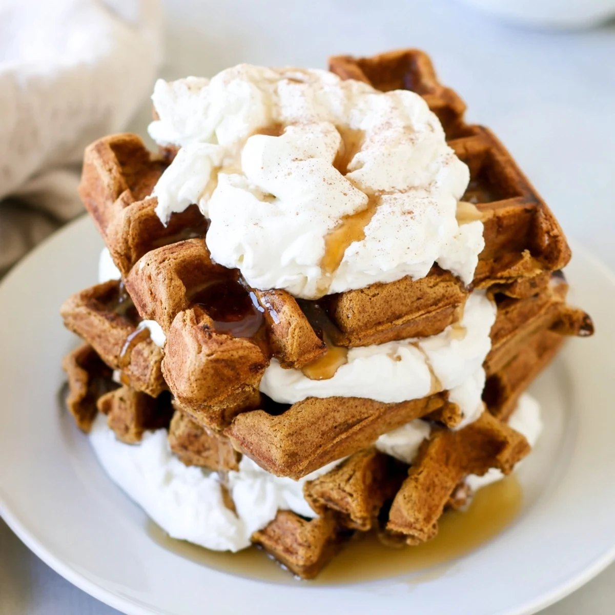 Warm gingerbread waffle stacks drizzled with maple syrup and fluffy whipped cream, tempting viewers.
