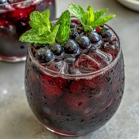 A glass of Sparkling Black Currant Mocktail with fresh berries and mint, poured over ice for a refreshing summer beverage.