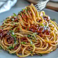 A steaming bowl of gochujang butter pasta glistens with glossy, spicy-sweet sauce, topped with sliced green onions and sesame seeds.